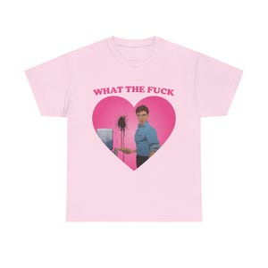 May include: Pale pink t-shirt featuring the text "WHAT THE FUCK" in pink above a heart-shaped graphic. The graphic shows an illustration of a person in a blue shirt and black trousers, with a water cooler and black splatters.
