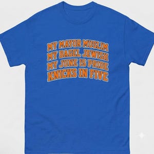 Op de afbeelding: Een koningsblauw t-shirt met korte mouwen en oranje tekst met een witte omlijning die luidt: MY MAYOR MUSLIM, MY BAGEL JEWISH, MY JUNE IS PRIDE, KNICKS IN FIVE. Het shirt heeft een ronde hals en een standaard pasvorm met vier tekstregels.