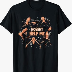 May include: Black t-shirt featuring a humorous graphic with the text "ROBERT HELP ME." The design includes several stylized insect-like creatures with speech bubbles and star accents. The creatures are in shades of brown and tan.