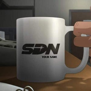 May include: A white ceramic mug with a black logo that reads "SDN" and "YOUR NAME" underneath. The mug is being held by a person's hand. The mug is on a wooden surface.