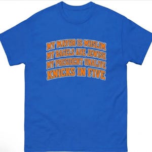 May include: A royal blue short-sleeve t-shirt featuring orange text with a white outline that reads: MY MAYOR IS MUSLIM, MY BAGELS ARE JEWISH, MY PRESIDENT UNALIVE, KNICKS IN FIVE. The text is arranged in four arched lines across the chest area.