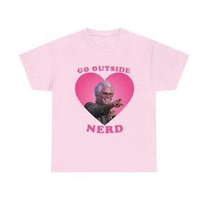 Pode incluir: Camiseta rosa com um gráfico de coração rosa que apresenta um homem com cabelo branco e barba. O texto "GO OUTSIDE NER" está impresso na camiseta.