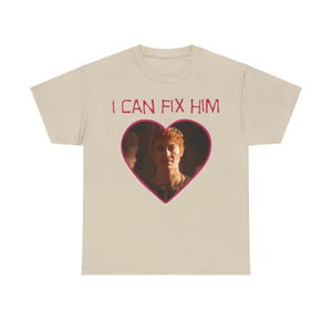 Puede incluir: Una camiseta beige con el texto "I CAN FIX HIM" impreso en rosa. Una imagen en forma de corazón de un hombre con cabello rojo está centrada debajo del texto.
