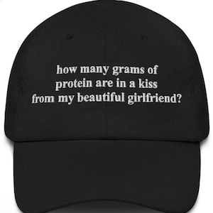 May include: Black baseball cap with white embroidered text: "how many grams of protein are in a kiss from my beautiful girlfriend?" The cap has a curved brim and a top button.