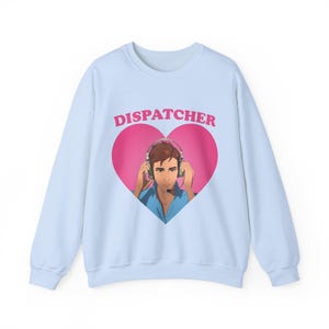 May include: Light blue sweatshirt with a pink heart graphic. The heart contains an illustration of a person wearing a headset. The word "DISPATCHER" is printed above the heart in pink. The sweatshirt has long sleeves and a crew neck.
