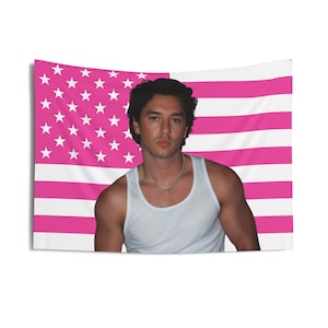 May include: A tapestry featuring a pink and white American flag design. A person wearing a white tank top and a silver chain necklace is centered in front of the flag. The person has dark hair and is looking at the viewer.
