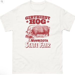 May include: White t-shirt with a red graphic. The graphic features two pigs and the text "Girthiest Hog" above and "At The Minnesota State Fair" below. The shirt has a crew neck.