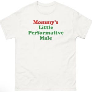 Puede incluir: Camiseta blanca con el texto "Mommy's Little Performative Male" en letras rojas y verdes. La camiseta tiene cuello redondo y mangas cortas. El texto está centrado en la parte delantera.