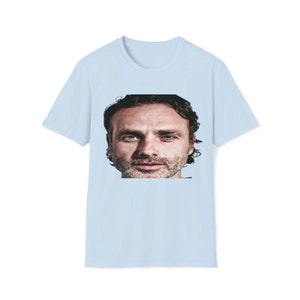 Può includere: Una t-shirt blu chiaro con un'immagine stampata di un viso maschile. L'uomo ha i capelli corti castani, la barba e guarda direttamente la telecamera.