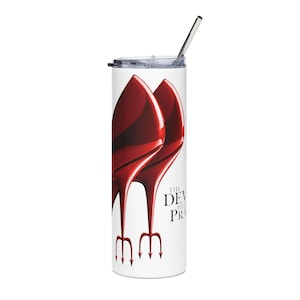 May include: A white tumbler featuring a red high heel design and the text "THE DEVIL WEARS PRADA". It has a silver straw and a clear lid. The design includes a pair of red high heels and pitchforks.
