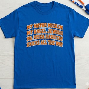 Op de afbeelding: Een koningsblauw T-shirt met korte mouwen en oranje tekst op de borst met de tekst: MY MAYOR MUSLIM, MY BAGEL JEWISH, MA RISKA HARGITAY, KNICKS ALL THE WAY. Het shirt heeft een ronde hals en een standaard pasvorm met een eenvoudig ontwerp.