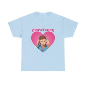 May include: Pale blue crew neck t-shirt with a pink heart design. The heart contains the word "DISPATCHER" above an illustration of a person wearing a headset. The person is wearing a blue shirt.