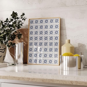 May include: A framed print with a white background and a repeating blue and white pattern of stylized flowers. The print is sitting on a countertop with a vase, a pitcher, and a bowl of lemons.