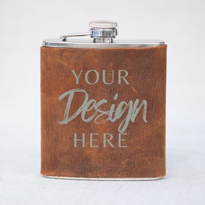 Puede incluir: Una petaca de acero inoxidable envuelta en cuero marrón con una tapa plateada. La petaca tiene el texto "YOUR Design HERE" grabado en la parte delantera.
