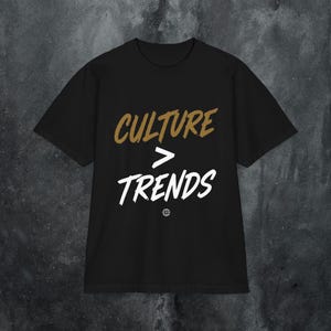 Puede incluir: Una camiseta negra de manga corta con el texto CULTURE en dorado, un símbolo de mayor que y la palabra TRENDS en blanco. Debajo del texto hay un pequeño icono de un globo terráqueo blanco. La camiseta tiene cuello redondo y corte estándar.