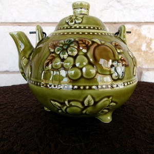 May include: A green ceramic teapot with a floral and fruit design. The teapot has a lid and a spout, and it is decorated with raised flowers and fruit.