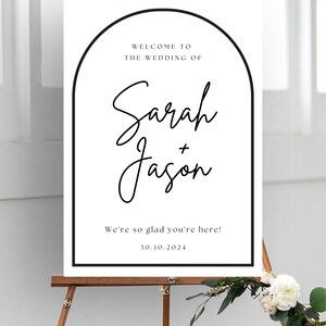 Op de afbeelding: Een wit welkomstbord voor een bruiloft op een houten ezel. Het bord heeft een zwarte boogvormige rand en de tekst "Welkom op de bruiloft van Sarah + Jason" en "We zijn zo blij dat je er bent!" met de datum 30.10.2024.