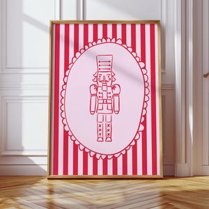 May include: A framed print featuring a red and white striped background. A pink oval in the center contains a red outline illustration of a nutcracker soldier. The frame is gold, and the print is displayed against a white wall and wooden floor.