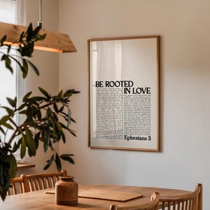 May include: A framed print with the words "BE ROOTED IN LOVE" and text from Ephesians 3. The print has a light-colored background and a wooden frame. The print is hanging on a white wall.