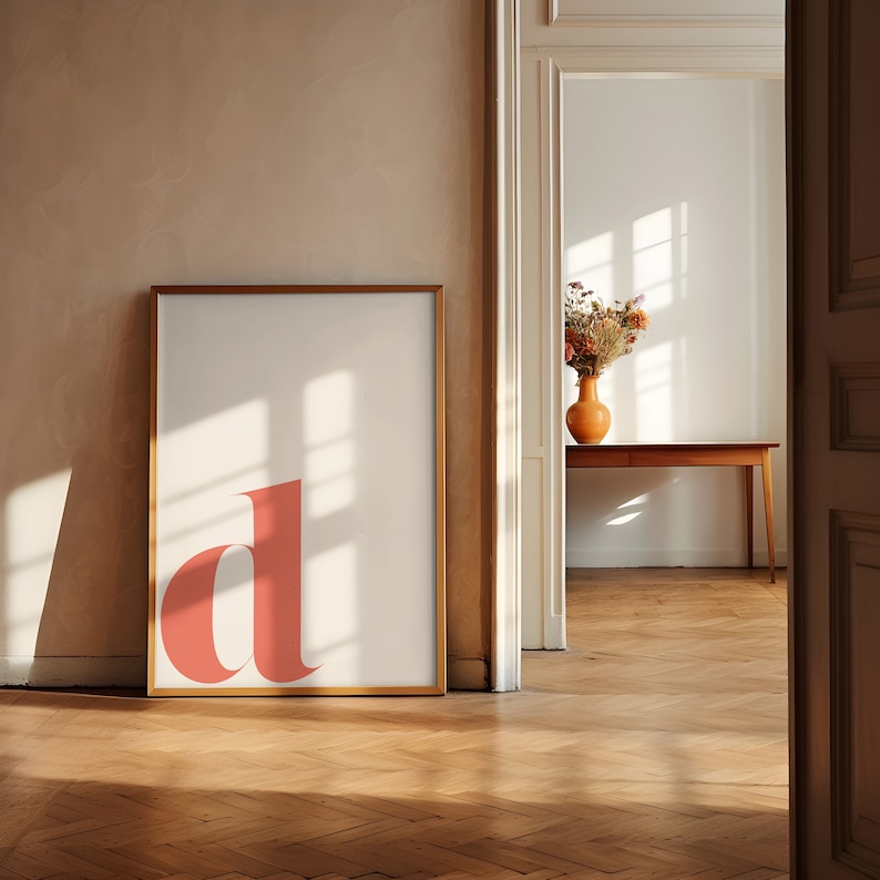 Pu&ograve; includere: Una stampa incorniciata con una grande lettera "d" di colore corallo in grassetto su sfondo bianco. La stampa &egrave; appesa a una parete color beige chiaro in una stanza con pavimento in legno. La luce del sole entra da una porta sullo sfondo, illuminando la stanza.