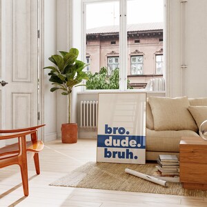 May include: A white poster with blue text that reads "bro. dude. bruh." The poster is hanging on a wall in a living room with a couch, a plant, and a chair.