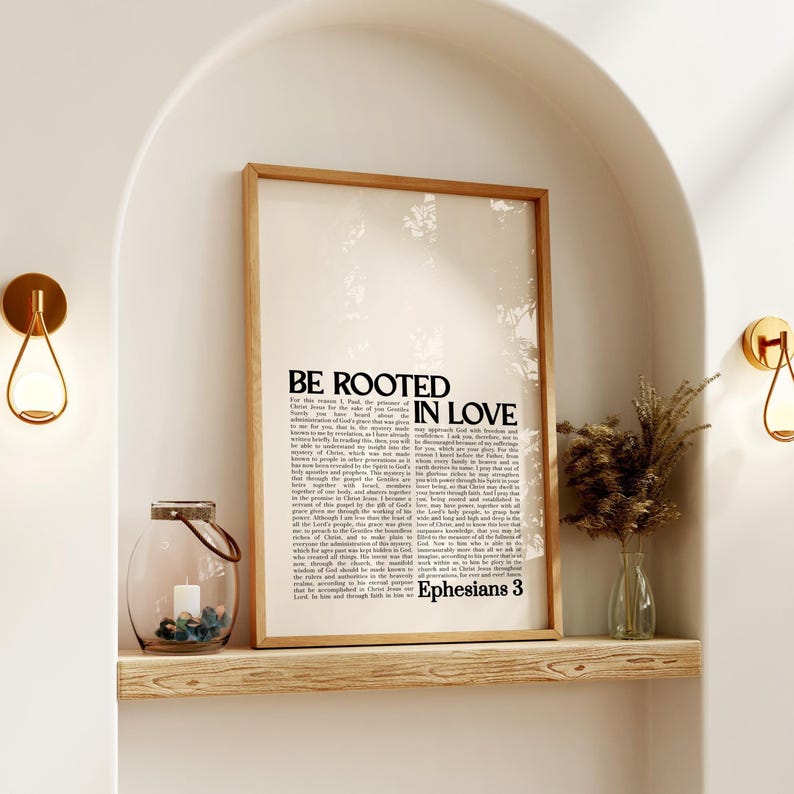 May include: A framed print with the words "BE ROOTED IN LOVE" and text from Ephesians 3. The print is in a light wood frame, displayed on a wooden shelf with a candle lantern and a small vase with dried plants. Two gold sconces flank the shelf.