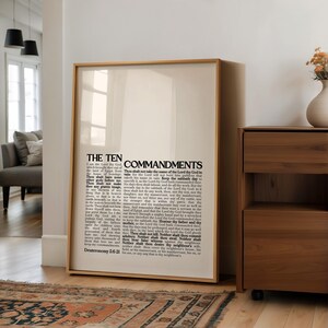 May include: A framed print featuring the text "THE TEN COMMANDMENTS" in a bold, black font. The text is centered on a cream-colored background, encased in a light brown wooden frame. The print is displayed against a white wall, with a glimpse of a living room setting.