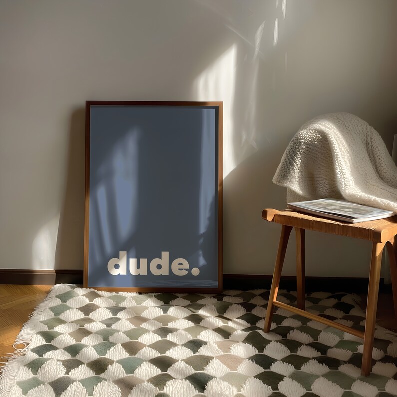 May include: A framed print with a blue background and the word "dude." in white letters. The print is sitting on a white and green patterned rug in front of a white wall.