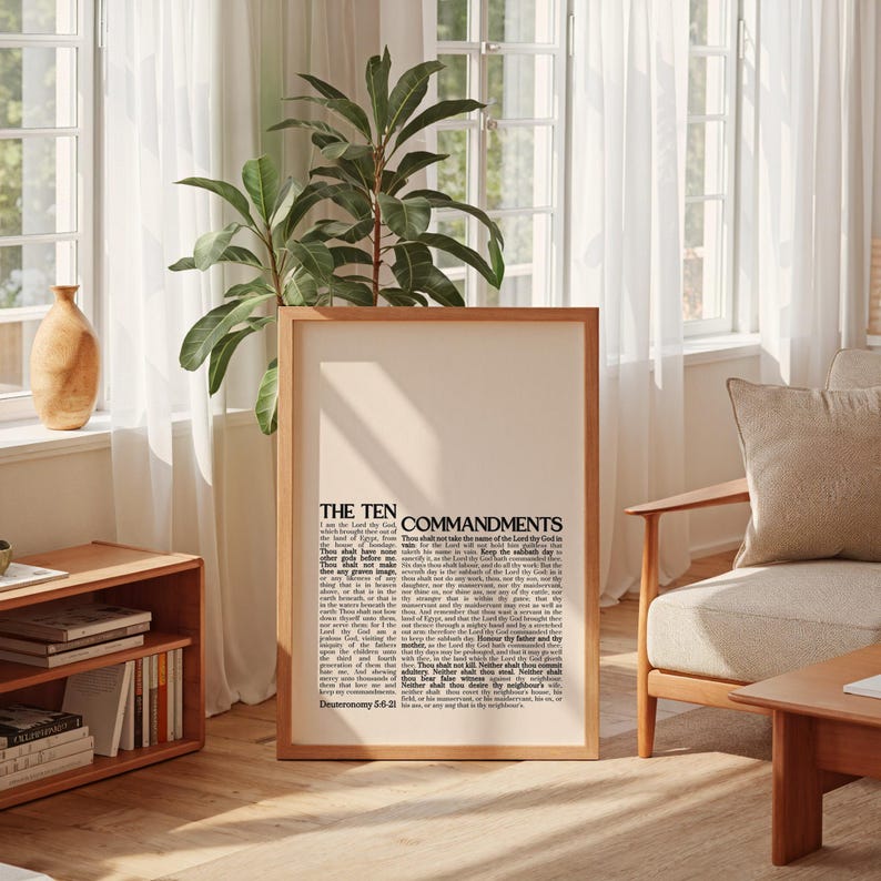 May include: A framed print featuring the text "THE TEN COMMANDMENTS" in a classic serif font. The print has a cream-colored background and a wooden frame. The image is displayed in a well-lit room with a plant and wooden furniture.