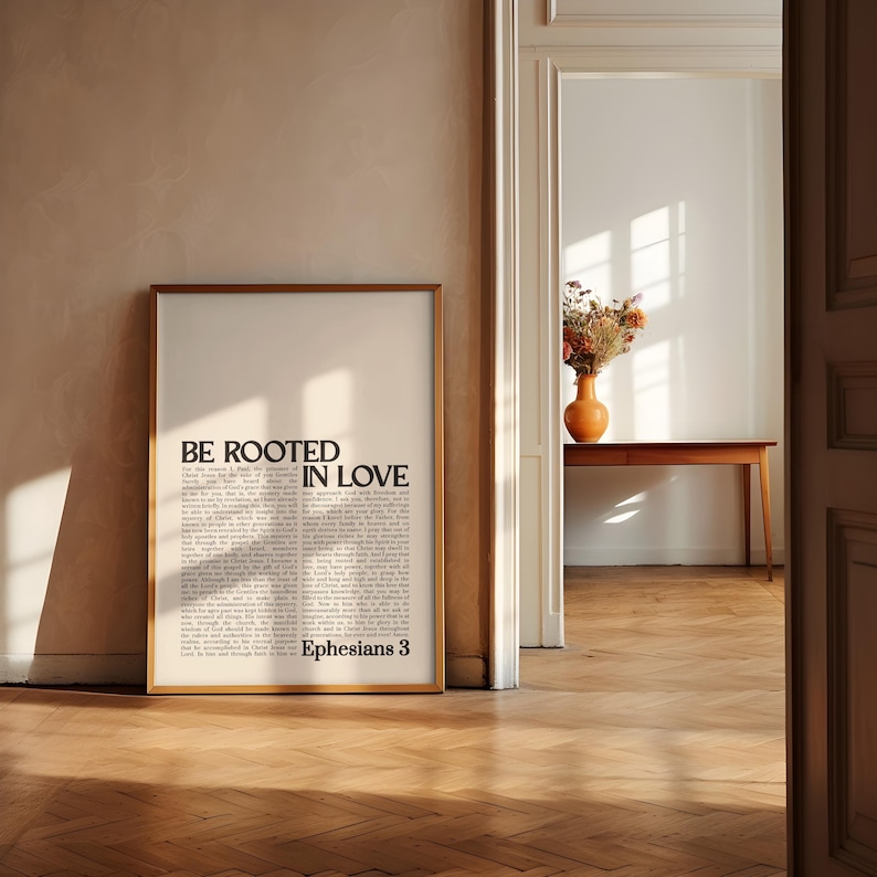 May include: A framed print with the words "BE ROOTED IN LOVE" in bold, black text. The print also includes a passage of text and the reference "Ephesians 3." The frame is a light brown color, and the background is a neutral beige.