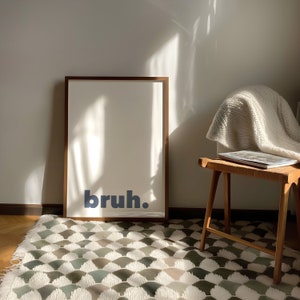May include: A framed print with the word "bruh" in blue text on a white background. The print is hanging on a white wall above a white rug with a geometric pattern.