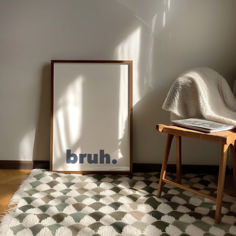 May include: A framed print with the word "bruh" in blue text on a white background. The print is hanging on a white wall above a white rug with a geometric pattern.