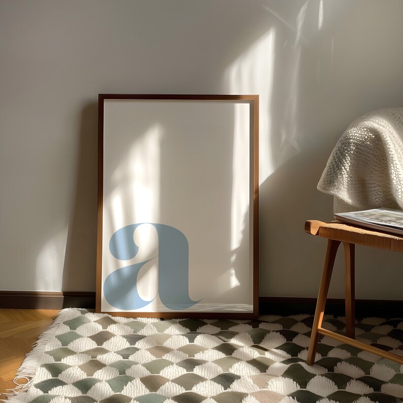 Pu&ograve; includere: Una stampa incorniciata con una lettera stilizzata "a" blu chiaro su sfondo bianco. La cornice &egrave; in legno marrone e la stampa &egrave; appesa a una parete bianca.