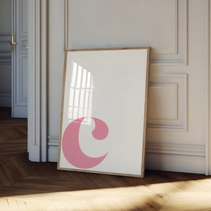 Pu&ograve; includere: Una stampa incorniciata con una lettera "C" rosa su sfondo bianco. La cornice &egrave; in legno e ha una finitura naturale.