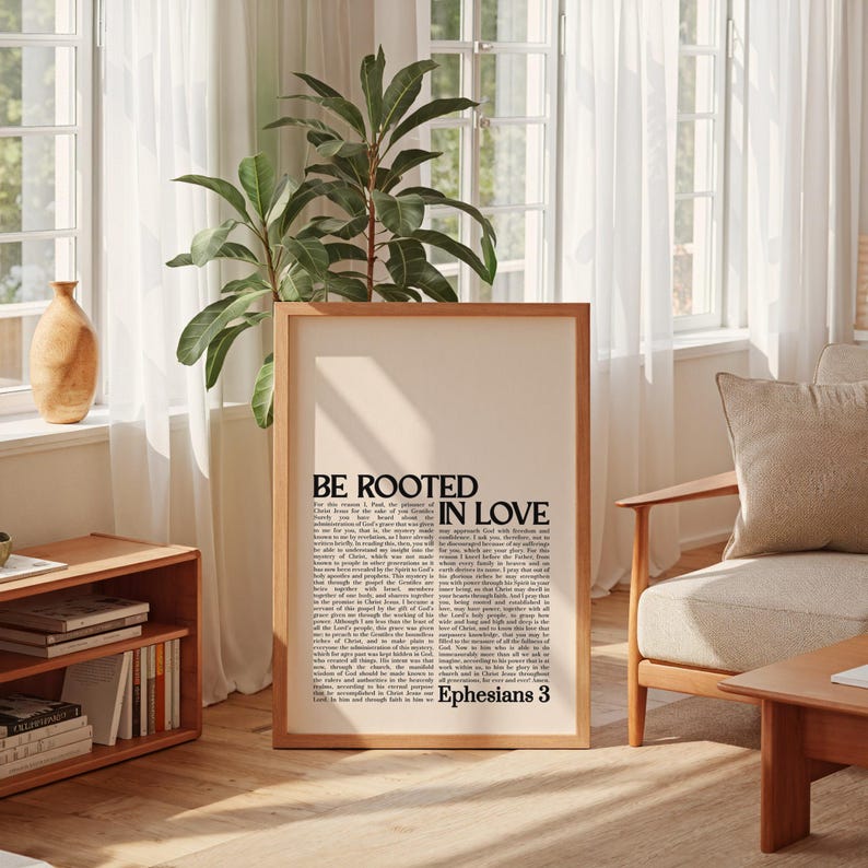 May include: A framed print with the text "BE ROOTED IN LOVE" and "Ephesians 3" in black font. The print is in a light wood frame and is displayed in a well-lit room with a plant and wooden furniture. The print has a cream-colored background.
