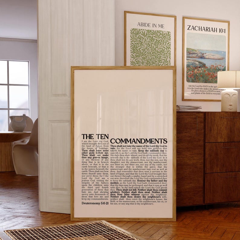 May include: A framed print of the Ten Commandments, with the text in black. Two other framed prints are visible, one with green foliage and the other with a coastal landscape. The prints are displayed in a room with wooden floors.