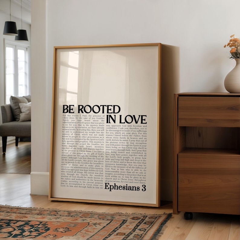 May include: A framed print with the words "BE ROOTED IN LOVE" in bold black letters. The print also contains a passage from Ephesians 3. The frame is a light brown color, and the print is displayed in a room with a wooden cabinet and a patterned rug.