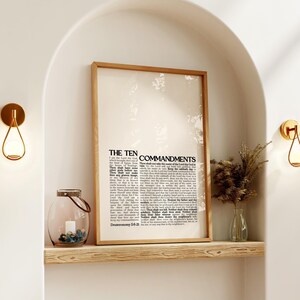 May include: A framed print of "The Ten Commandments" with a light wood frame. The print is displayed on a wooden shelf, alongside a glass lantern with stones and a small vase with dried plants. Two gold sconces are mounted on the wall.