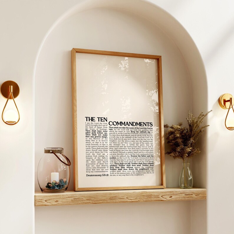 May include: A framed print of "The Ten Commandments" with a light wood frame. The print is displayed on a wooden shelf, alongside a glass lantern with stones and a small vase with dried plants. Two gold sconces are mounted on the wall.