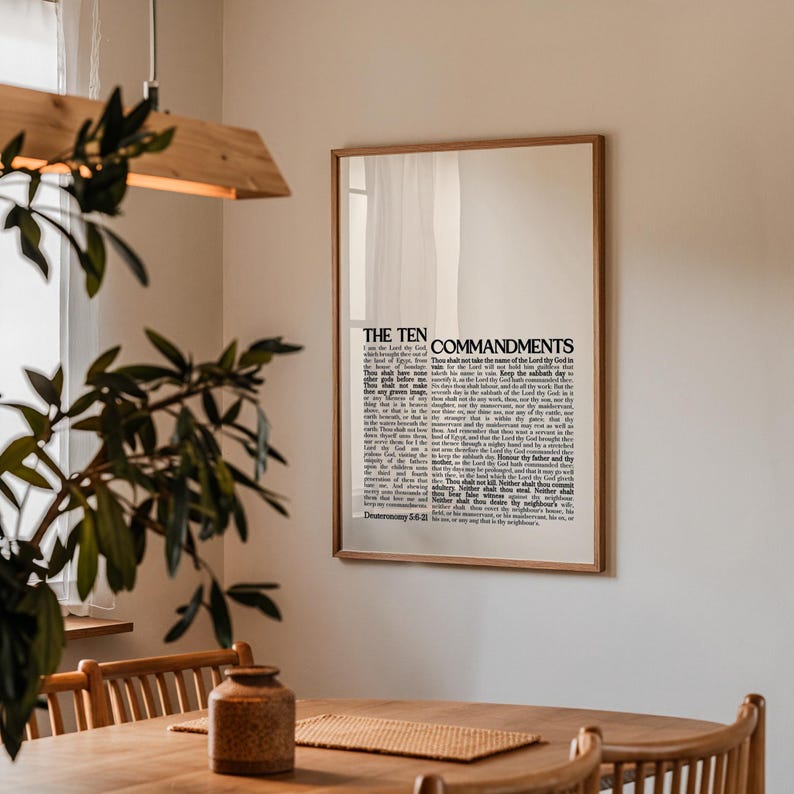 May include: A framed print featuring the text "The Ten Commandments" in a classic serif font. The print is in a wooden frame and hangs on a white wall. The image is in a well-lit room with a wooden table and a plant.