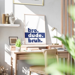 May include: A white framed poster with the text "bro. dude. bruh." in blue lettering on a white background. The poster is hanging on a wall above a wooden desk with a lamp and a potted plant.