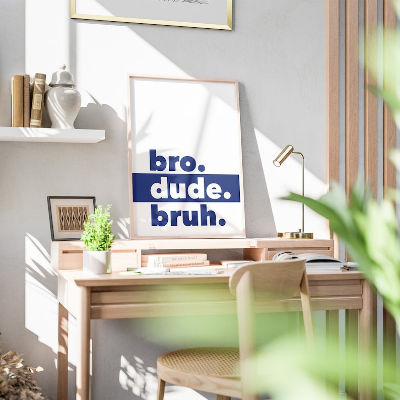 May include: A white framed poster with the text "bro. dude. bruh." in blue lettering on a white background. The poster is hanging on a wall above a wooden desk with a lamp and a potted plant.