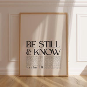 May include: A framed print featuring the words "BE STILL & KNOW" in large, bold, black lettering. The scripture Psalm 46 is below the text. The background is a light beige, and the frame is a light wood colour.