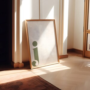 Pu&ograve; includere: Un'immagine incorniciata in oro con uno sfondo bianco e una lettera 'j' verde al centro. L'immagine &egrave; appoggiata contro un muro bianco.