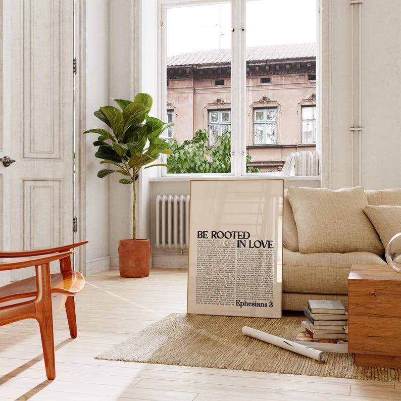 May include: A framed poster with the text "BE ROOTED IN LOVE" and "Ephesians 3" in black font. The poster is leaning against a beige sofa in a well-lit room with a large window and a potted plant.