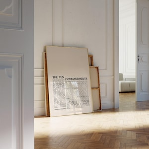 May include: A framed print of "The Ten Commandments" in a minimalist style. The text is in black on a cream-colored background, with a wooden frame. The print is leaning against a wall in a room with a wooden floor.
