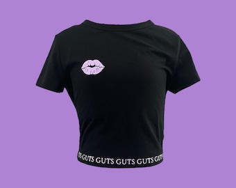 May include: Black cropped t-shirt with a purple lips graphic and the text "GUTS GUTS GUTS GUTS GUTS" printed on the bottom hem.