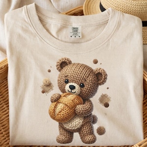 May include: A beige Comfort Colors t-shirt featuring a knitted teddy bear holding a ball of yarn. The bear is brown and cream-colored, with a black nose and eyes. The shirt is displayed in a woven basket.