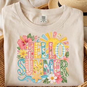May include: Beige t-shirt with a colorful "Hello Summer" graphic. The design features floral elements, a sun, and a starfish. The shirt is folded in a wicker basket, with a straw hat in the background.