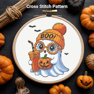 May include: Cross-stitch pattern featuring a cute ghost wearing an orange beanie with "BOO!" on it, round glasses, and holding a pumpkin and a drink. The design is set in a wooden embroidery hoop, with pumpkins and bats around it.
